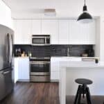 Zero Dry Time Modern kitchen with stainless steel appliances, white cabinets, black backsplash, and a white island with two black stools under pendant lights. A framed picture hangs on the wall.
