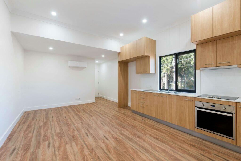 Zero Dry Time Modern, unfurnished kitchen and living area with wood flooring, wood cabinets, built-in oven, stovetop, window, and recessed ceiling lights.